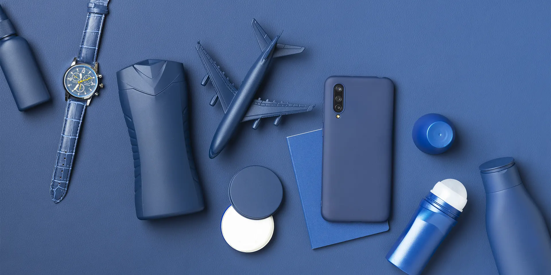 Composição de itens executivos azuis sobre fundo azul, destacando relógio de pulso, miniatura de avião, smartphone, frascos e acessórios de viagem da LCM Brindes.