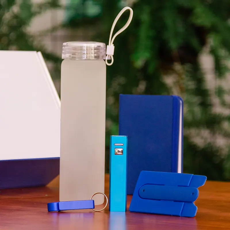 Kit de brindes personalizados contendo garrafa de vidro fosco, bloco de notas azul, power bank, abridor de garrafas e suporte para celular sobre mesa de madeira.